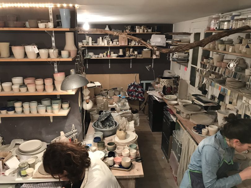 Atelier de poterie. Salle remplie de vases, verres et assiettes sur les étagères. On peut voir deux femmes travailler dans les coins gauche et droite du bas de l'image