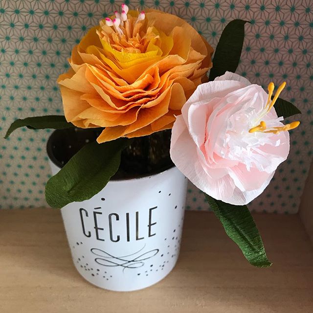 Fleurs en papier fabriquées dans un atelier Make my lemonade marque de vêtements créée par Lisa Gachet. Il y a une fleur orange et une fleur blanche en papier dans un pot avec écrit dessus Cécile. En arrière plan un fond avec motifs.