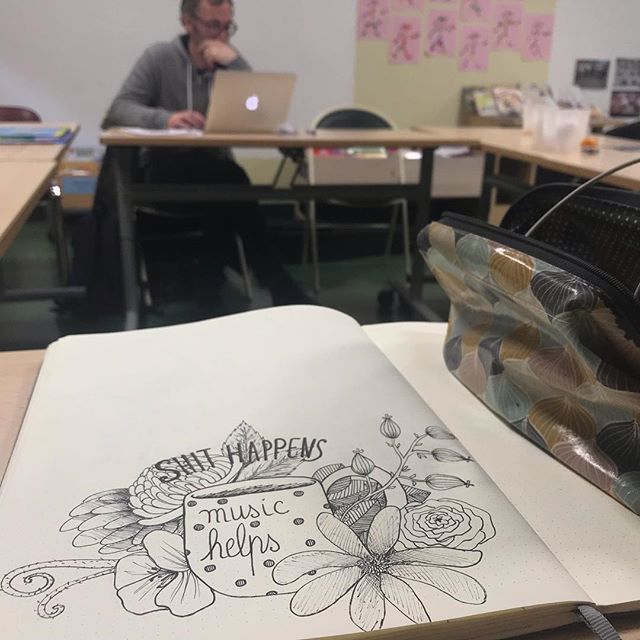 Dessin d'un mug avec des plantes, fleurs à l'arrière. Dessin sur un cahier. Derrière en arrière plan on peut voir un homme travailler sur son Mac. C'est Olivier du groupe de musique Volo