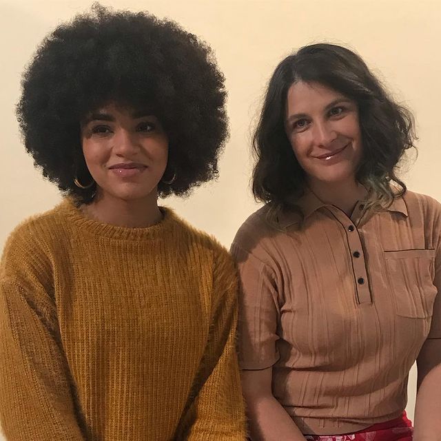 2 femmes qui sourient pour un shooting photo. Une d'entre elle est Ciloubidouille. L'autre a une afro, de gros boucles d'oreilles dorés, et un pull orange. 